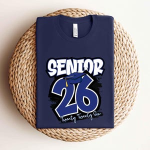 May include: Navy blue t-shirt with the text "SENIOR 26" in white and blue. The design includes a graduation cap and the words "Twenty Twenty Six" in white. The shirt is displayed on a woven, circular surface.