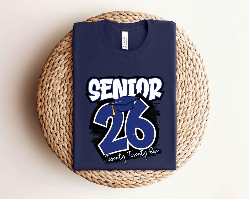 May include: Navy blue t-shirt with the text "SENIOR 26" in white and blue. The design includes a graduation cap and the words "Twenty Twenty Six" in white. The shirt is displayed on a woven, circular surface.
