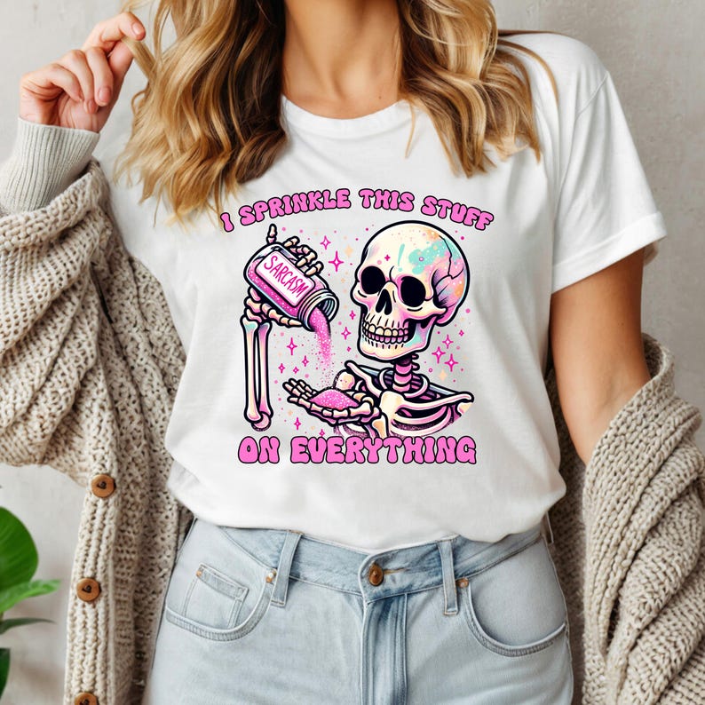 May include: White t-shirt with a colorful skeleton graphic. The skeleton is pouring pink glitter from a container labeled "Sarcasm." The text "I Sprinkle This Stuff On Everything" is above and below the graphic.