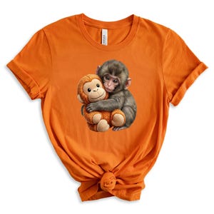 May include: Orange t-shirt featuring a graphic of two monkeys hugging. One monkey is gray and the other is a plush toy in shades of brown and tan. The t-shirt is made of a soft material.