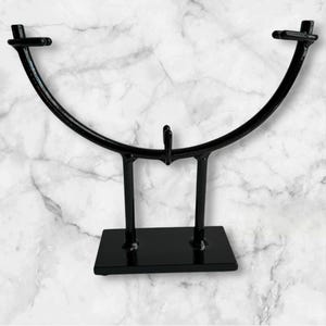 May include: A black metal display stand with a curved top and a rectangular base. The stand has a minimalist design, with two vertical supports connecting the base to the curved top. The stand is designed to hold an item.