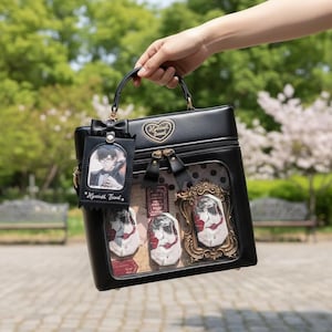 May include: Black handbag with a clear display window showcasing framed artwork and character pins. The bag features a top handle, a bow, and a character tag. The bag is held up against a blurred outdoor backdrop.