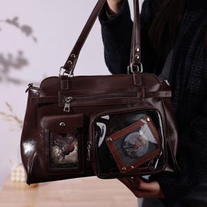 May include: A brown leather handbag with a shoulder strap and a smaller, detachable clear pouch. The handbag has a front pocket with a decorative image. The clear pouch has a brown frame and a circular image inside.