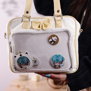 Peut inclure: Sacoche crème avec un nœud décoratif et une poche avant transparente. Le sac a des poignées et une bandoulière crème. Il est orné d'épingles et d'un porte-clés en forme de chat avec un personnage d'anime aux cheveux bleus.