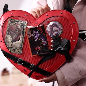 Może przedstawiać: Czerwona torebka w kształcie serca z czarnymi akcentami, rogami i skrzydłami nietoperza. Torba ma przezroczysty panel przedni z kartami tarota i guzikiem. Czarna kokarda i paski.