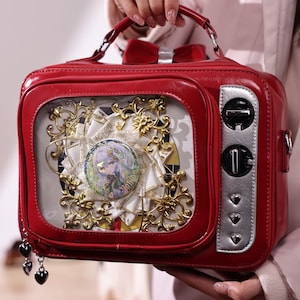 Peut inclure: Un sac à main rouge en forme de téléviseur vintage. Le sac présente un écran transparent affichant un arrangement décoratif avec un badge. Il possède une poignée, un nœud et des détails argentés. Un charm avec des pendentifs en forme de cœur est suspendu sur le côté.