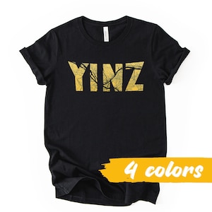 Peut inclure: T-shirt noir avec le mot "YINZ" dans un motif de carte doré. Le t-shirt a des manches retroussées et une bannière jaune en bas avec l'inscription "4 couleurs".