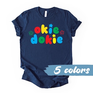 May include: Navy blue t-shirt with the words "okie dokie" in colorful bubble letters. The design includes a flower, star, and other decorative elements. The text "5 colors" is displayed in a light blue banner.