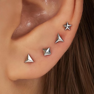 Può includere: Primo piano di un orecchio adornato da cinque orecchini a lobo argentati. Gli orecchini includono un triangolo, un quadrato, una piramide e una stella. L'orecchio è posto su uno sfondo sfocato di capelli castani.