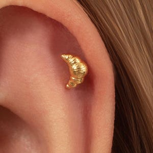 May include: A small gold earring shaped like a croissant, featuring a textured surface with curved ridges. The piece is positioned in the upper cartilage area of an ear, showcasing a metallic finish and a detailed, realistic pastry design.