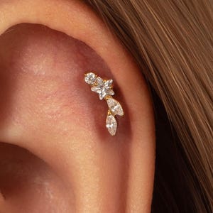Könnte beinhalten: Ein goldfarbener Knorpel-Ohrring mit einer Anordnung aus klaren, facettierten Steinen. Das Design besteht aus einem kleinen sternförmigen Stein an der Spitze, gefolgt von zwei marquise-geschliffenen Steinen in einer vertikalen, blattartigen Form.