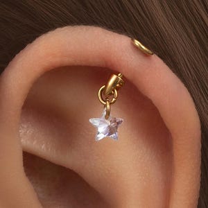 May include: A gold helix earring featuring a small, clear star-shaped charm. The earring is attached to the ear with a small hoop. A second gold hoop earring is also visible on the ear.