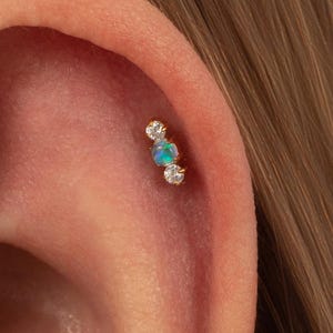 May include: A gold-toned cartilage earring featuring a central round iridescent opal flanked by two small clear round stones. The jewellery is positioned in the upper ear cartilage, showcasing a delicate three-stone linear design with prong settings.