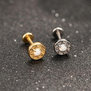 May include: Two flat-back stud earrings, one gold-toned and one silver-toned, each featuring a circular face with a sunburst pattern and a central white opal-like stone. These small jewellery pieces have a post back and a circular flat base.