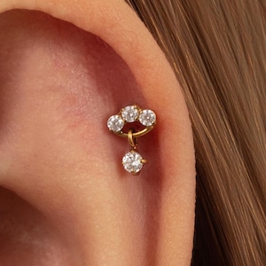 May include: A gold-toned cartilage earring featuring a curved bar set with three small clear round stones, with a single dangling clear round stone suspended from the centre, secured in the upper ear cartilage with a flat back post.