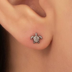 May include: A small, dark-toned metal stud earring shaped like a sea turtle. The turtle design features a textured shell with intricate detailing on the head and flippers, securely fastened to the earlobe as a single, compact piece of jewelry.