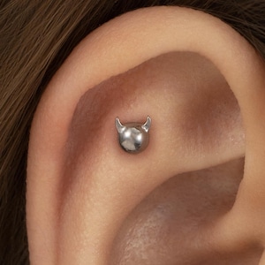 Puede incluir: Un pendiente de piercing para la oreja de metal plateado con forma de pequeña esfera lisa y dos cuernos puntiagudos en la parte superior. La joya está situada en la zona plana del cartílago de la oreja y tiene un acabado pulido y brillante.