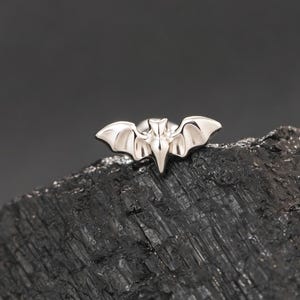 Può includere: Un piccolo orecchino a lobo in metallo color argento a forma di pipistrello stilizzato con ali spiegate. Il pipistrello presenta una superficie liscia e lucida con contorni definiti per la testa e le ali, creando una silhouette distinta.