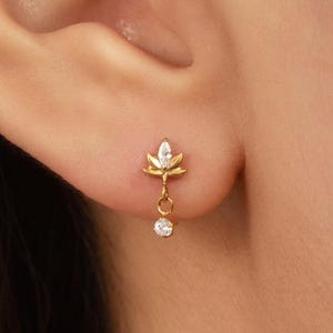 May include: A gold stud earring featuring a small, pointed leaf-shaped element set with a marquise-cut clear stone. A delicate gold chain hangs from the leaf, ending in a single, small, round, clear stone that drapes below the earlobe.