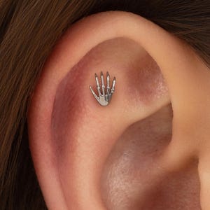 Könnte beinhalten: Ein silberfarbener Ohrstecker in Form einer Skeletthand mit fünf ausgeprägten Fingern. Das kleine, detaillierte Schmuckstück ist im oberen Knorpelbereich eines Ohrs platziert und besitzt eine polierte Oberfläche, die das Licht reflektiert.