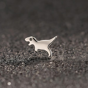 Könnte beinhalten: Ein kleiner, silberfarbener Ohrstecker in Form eines Dinosauriers. Der Ohrring hat eine glatte, polierte Oberfläche und ein minimalistisches Design. Der Dinosaurier ist im Profil dargestellt, mit offenem Maul und erhobenem Schwanz. Der Ohrring ist vor einem dunklen, strukturierten Hintergrund platziert.
