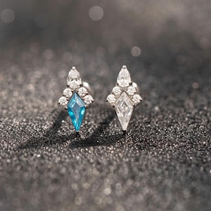 May include: A pair of stud earrings featuring a kite-shaped design. One earring contains a central blue gemstone, while the other features a clear gemstone. Both are accented with small, round, clear stones arranged in a cluster at the top.