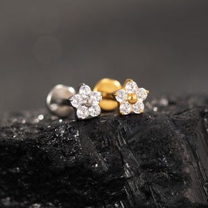 May include: Close-up of two flower-shaped stud earrings. One earring is silver-toned with clear stones, the other is gold-toned with clear stones. The earrings are set on a dark, textured surface.