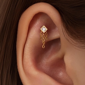 Può includere: Primo piano di un orecchino color oro con una piccola gemma trasparente e quadrata nella parte superiore. L'orecchino ha un design a catena pendente con più piccoli anelli circolari. L'orecchino è posizionato sull'orecchio.