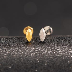 Könnte beinhalten: Zwei Ohrstecker mit einem marquiseförmigen Design. Einer ist goldfarben, der andere silberfarben. Sie sind vor einem dunklen, strukturierten Hintergrund platziert.