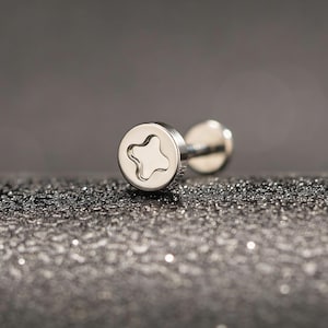 Può includere: Un orecchino a lobo color argento con una faccia circolare con un design a forma di stella. L'orecchino è posto su uno sfondo scuro e strutturato, creando un contrasto che ne evidenzia la lucentezza metallica. Un gioiello.