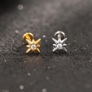 Puede incluir: Dos pendientes en forma de estrella, uno dorado y otro plateado, cada uno con una gema transparente en el centro. Se exhiben sobre una superficie oscura y texturizada, creando un contraste que resalta su diseño.