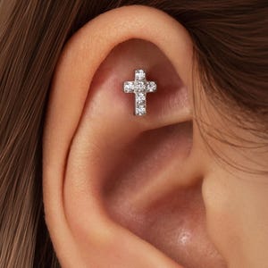 May include: A small, silver-toned cross earring with multiple clear, sparkling stones. The cross is set in a person's ear, showcasing the jewelry's design and placement. The earring is a religious symbol and a fashion accessory.
