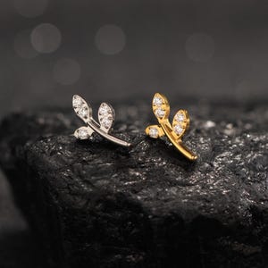 Peut inclure: Deux boucles d'oreilles en forme de feuille, une argentée et une dorée, chacune ornée de petits brillants étincelants. Les boucles d'oreilles sont posées sur un fond sombre et texturé, soulignant leur design délicat et leurs détails complexes. Ces boucles d'oreilles sont un accessoire élégant.