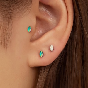 Könnte beinhalten: Nahaufnahme eines Ohrs mit drei kleinen Ohrringen. Zwei Ohrringe sind oval geformt und haben eine blau-grüne schillernde Farbe. Der dritte Ohrring ist ein kleiner, weißer, ovaler Stecker.