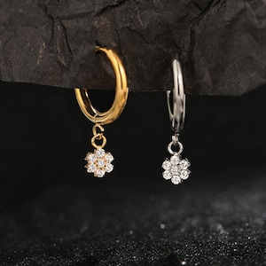Peut inclure: Deux boucles d'oreilles créoles, une dorée et une argentée, chacune ornée d'un petit pendentif en forme de fleur. Les pendentifs sont ornés de nombreuses pierres claires et scintillantes, pour un look délicat et élégant.