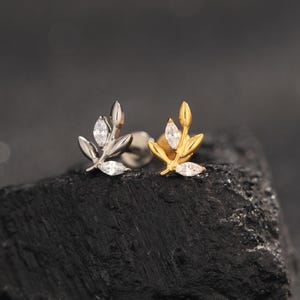 Peut inclure: Deux boucles d'oreilles en forme de feuille, une argentée et une dorée, chacune ornée de petites pierres précieuses transparentes. Les boucles d'oreilles sont présentées sur un fond sombre et texturé, soulignant leur design délicat et leur éclat.