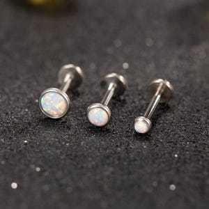 Può includere: Tre orecchini labret color argento con pietre iridescenti simili all'opale. Gli orecchini sono disposti in fila, con il più grande a sinistra e il più piccolo a destra. Lo sfondo è una superficie scura e strutturata.