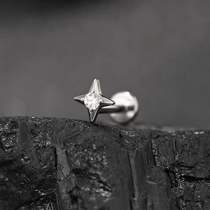 May include: A silver stud earring with a star-shaped design and a central clear gemstone. The earring is set against a dark, textured background. The design showcases the earring's reflective qualities and intricate details.