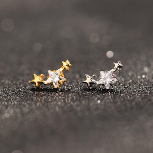 Peut inclure: Deux boucles d'oreilles en forme d'étoile. L'une est dorée, l'autre argentée. Chaque boucle d'oreille présente un groupe d'étoiles, avec une étoile en cristal clair plus grande au centre du design. Les boucles d'oreilles sont posées sur un fond sombre et scintillant.