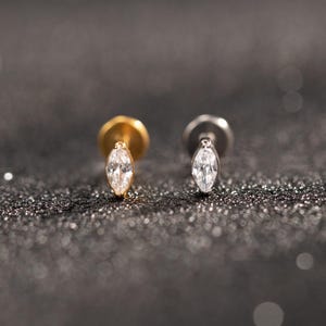 Könnte beinhalten: Zwei Ohrstecker mit einem klaren, marquiseförmigen Edelstein. Einer hat einen goldfarbenen Stift und Verschluss, der andere einen silberfarbenen Stift und Verschluss. Sie sind vor einem dunklen, glitzernden Hintergrund platziert.