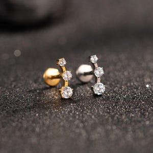 May include: Two stud earrings, one gold-coloured and one silver-coloured, each with three clear, sparkling round-cut stones in a vertical line. The earrings are set against a dark, textured background, contrasting their brilliance.