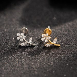 May include: Two flower-shaped stud earrings. One is silver-toned, the other gold-toned. Each earring features a cluster of small, clear gemstones forming the flower's center, with gemstone leaves. The earrings are set against a dark, textured background.