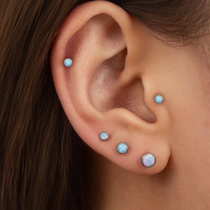 May include: Close-up of an ear with multiple opal stud earrings. The earrings are round with a light blue and white iridescent color. The ear has five earrings in various sizes.