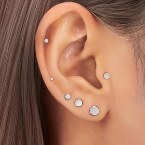 Peut inclure: Gros plan sur une oreille ornée de plusieurs petites boucles d'oreilles rondes en argent. Les boucles d'oreilles, de tailles différentes, sont placées sur le cartilage et le lobe de l'oreille. Le teint est clair et les cheveux sont châtain clair.