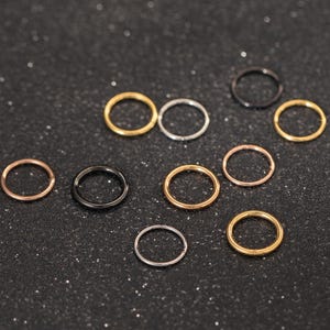 Peut inclure: Une collection de petits anneaux de nez circulaires dans différentes finitions métalliques, dont l'or, l'argent, l'or rose et le noir. Les anneaux sont disposés sur une surface sombre et scintillante, mettant en valeur leurs couleurs et styles.
