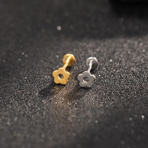 Puede incluir: Dos pendientes de botón con forma de flor. Uno es dorado y el otro plateado. Los pendientes tienen un pequeño diseño de flor de cinco pétalos. El fondo es una superficie oscura y texturizada.