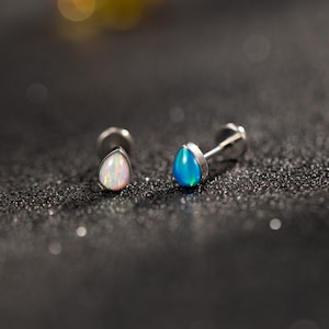 Può includere: Due orecchini a lobo in argento con pietre di opale a forma di goccia. Uno presenta un opale bianco con sfumature rosa, l'altro un opale blu brillante. Gli orecchini sono su uno sfondo scuro e strutturato.