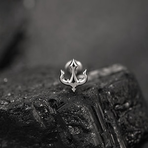 May include: A silver-toned metal stud earring shaped like a stylized anchor with a pointed top and curved flukes. The jewelry piece features a polished, reflective surface and is positioned upright, showcasing its symmetrical design and metallic finish.