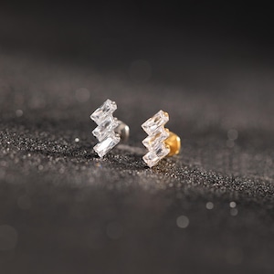 Può includere: Un paio di orecchini a bottone con un design a zig-zag. Un orecchino è argentato e l'altro dorato. Ogni orecchino presenta tre cristalli rettangolari trasparenti. Gli orecchini sono esposti su una superficie scura e strutturata.