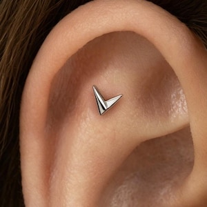 Peut inclure: Une boucle d'oreille en métal argenté en forme de petit chevron en V pointu. Le bijou est placé dans la zone du cartilage supérieur de l'oreille, présentant une surface polie et réfléchissante avec des lignes nettes et une pointe vers le bas.